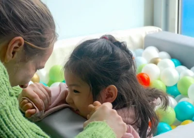 An autistic little girl playing in a ball pit with her therapist. Both of them are looking at each other and smiling