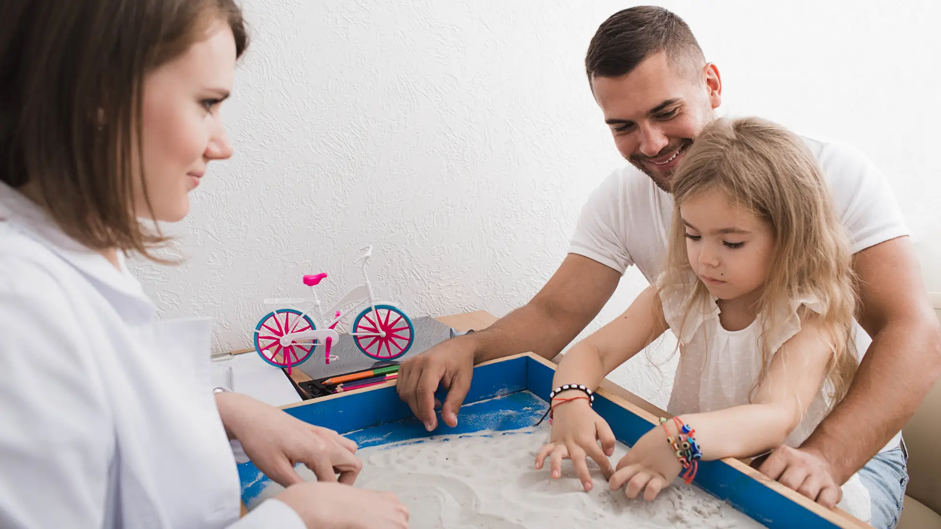 An autistic little girl with her dad and a therapist playing with sand and looking happy