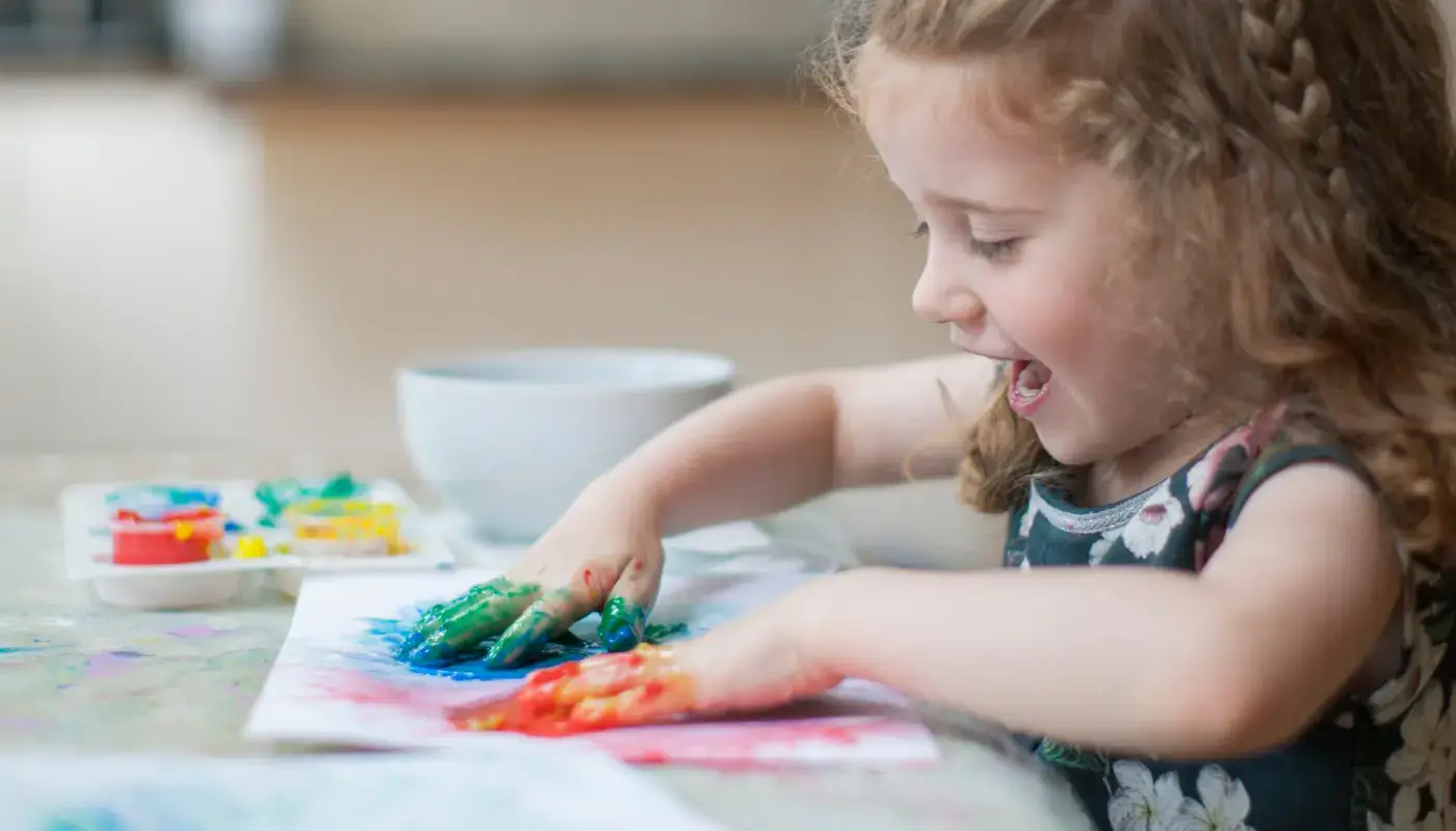 A little girl having fun doing finger painting on a piece of paper. Her hands are full of red and blue paint