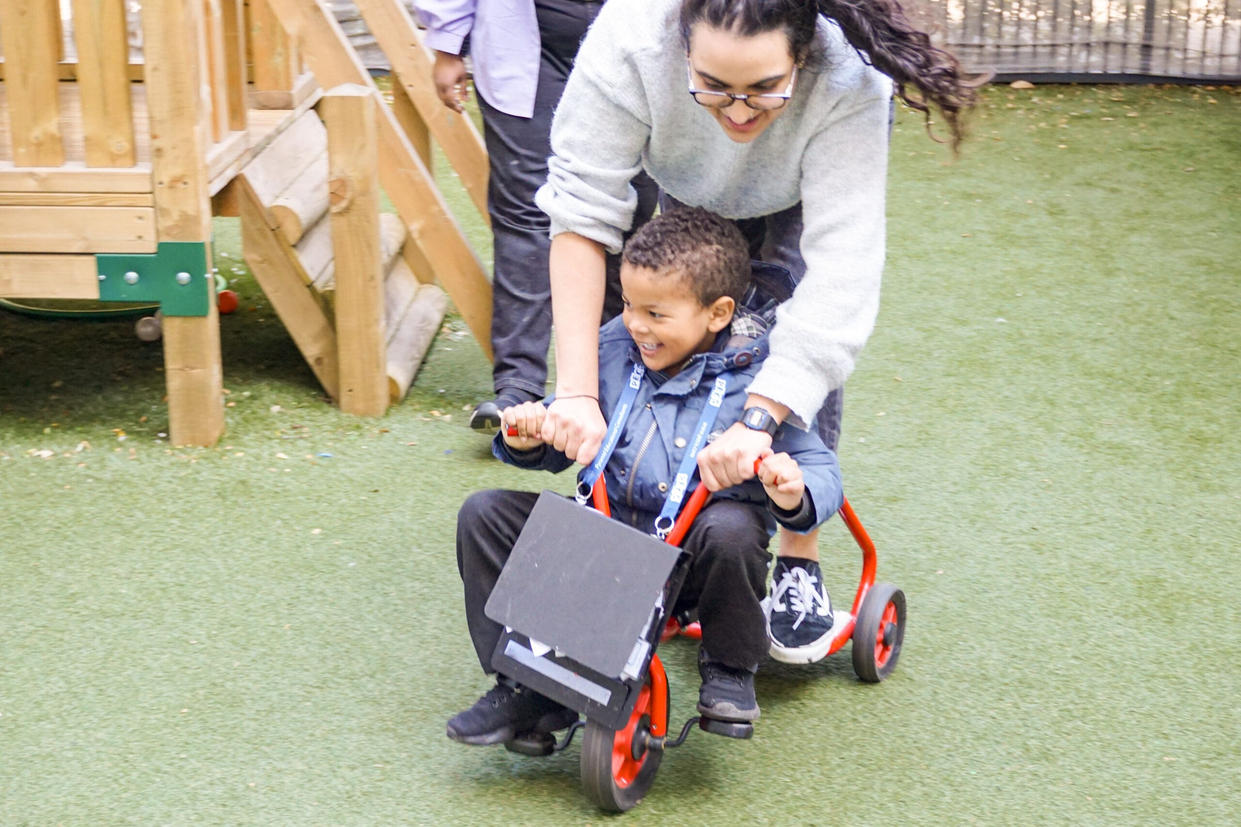 An autistic child smiles with excitement as they ride a tricycle, guided by a supportive ABA therapist in an outdoor play area. The moment captures the joyful, confidence-building experiences children enjoy at First Bridge Centre’s summer school where learning through movement, play, and encouragement takes centre stage.