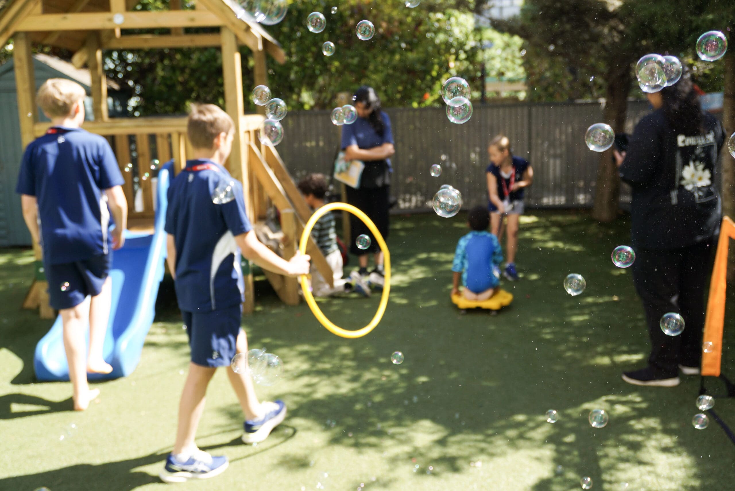 Children from Danes Hill School join a therapeutic play session at First Bridge Education, engaging in inclusive outdoor activities with bubbles, group games, and a climbing frame. The image captures the joyful, community-led atmosphere that supports social development and early intervention for children with additional needs.