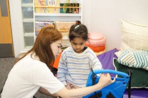 A young child at First Bridge School sits on a padded mat while an adult supports her in using a blue AAC (Augmentative and Alternative Communication) device. The adult is guiding her hand or showing her how to use the device. Behind them is a bookshelf with children’s books and soft play cushions, indicating a supportive early years or therapy environment.