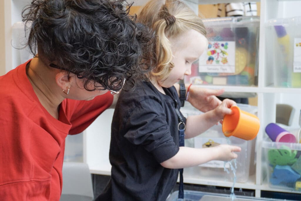 A young autistic child explores water play with an orange cup, supported by an adult at their side. They are engaged in a sensory-rich activity at First Bridge Education, surrounded by labelled storage boxes filled with toys. The environment is calm, structured, and child-centred—supporting the principles of routine, sensory regulation, and connection.