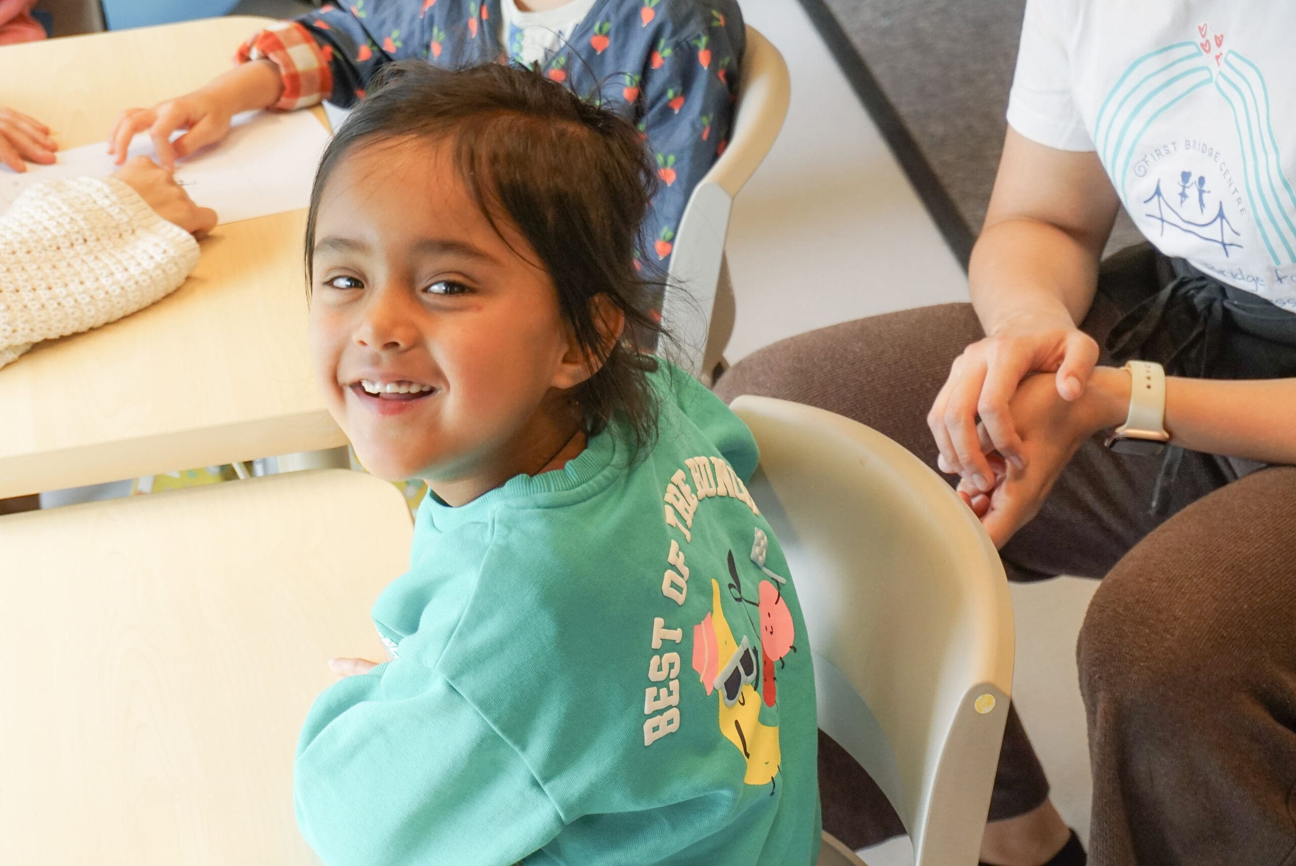 A child sits at a table in a SEN school classroom, smiling joyfully at the camera. Other children and a teacher are nearby, creating a warm and supportive learning environment.