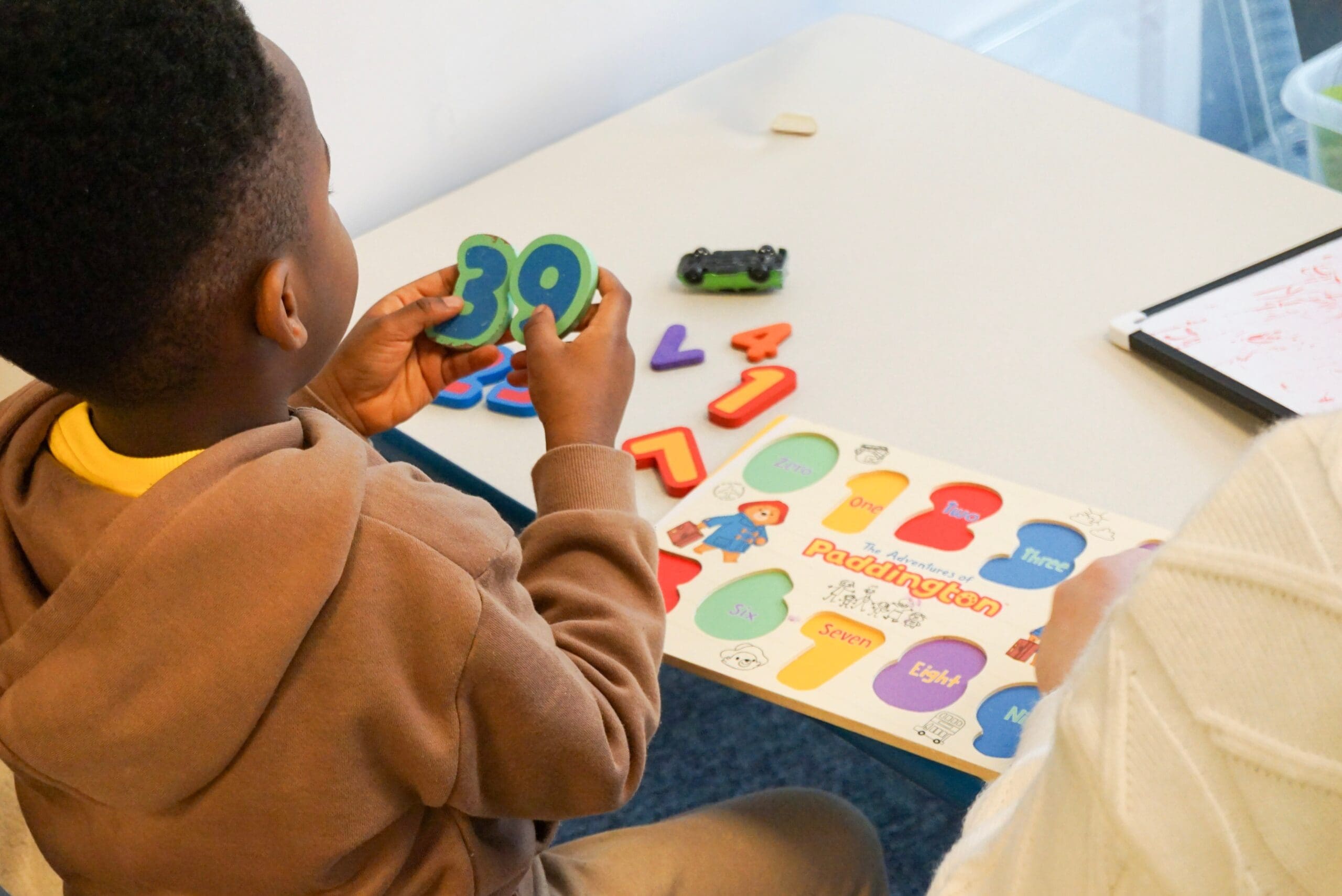 On the right, a young child sits at a table at First Bridge Education holding number shapes while working on a colourful learning activity.